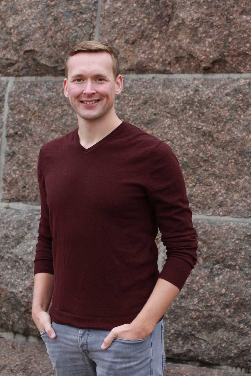 Zu sehen ist ein lächelnder Pascal Grüne in rotem Langarm-Shirt und Jeans stehend vor einer Steinwand.